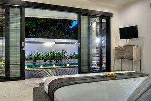 Quarto com cama, televisão e vista para uma piscina e área verde.