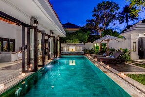 Uma área de piscina moderna com piscina transparente, espreguiçadeiras e um espaço de estar ao ar livre bem iluminado.
