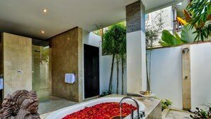 A modern bathroom with a large bathtub filled with rose petals, a statue, and a shower area. - The Residence Seminyak (Seminyak)
