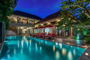 Piscina com espreguiçadeiras e guarda-sóis, cercada por um edifício com telhado de palha e deck de madeira.
