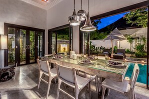 Uma área de refeições com mesa de vidro, cadeiras brancas e iluminação pendente.