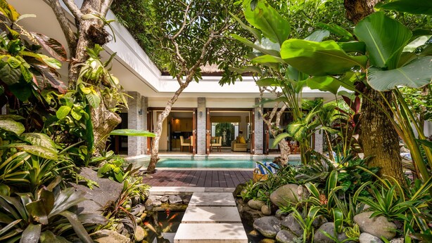 A pool area with a stone pathway leading to a pool, surrounded by lush tropical plants and a modern building with large glass doors. - The Residence Seminyak (Seminyak)