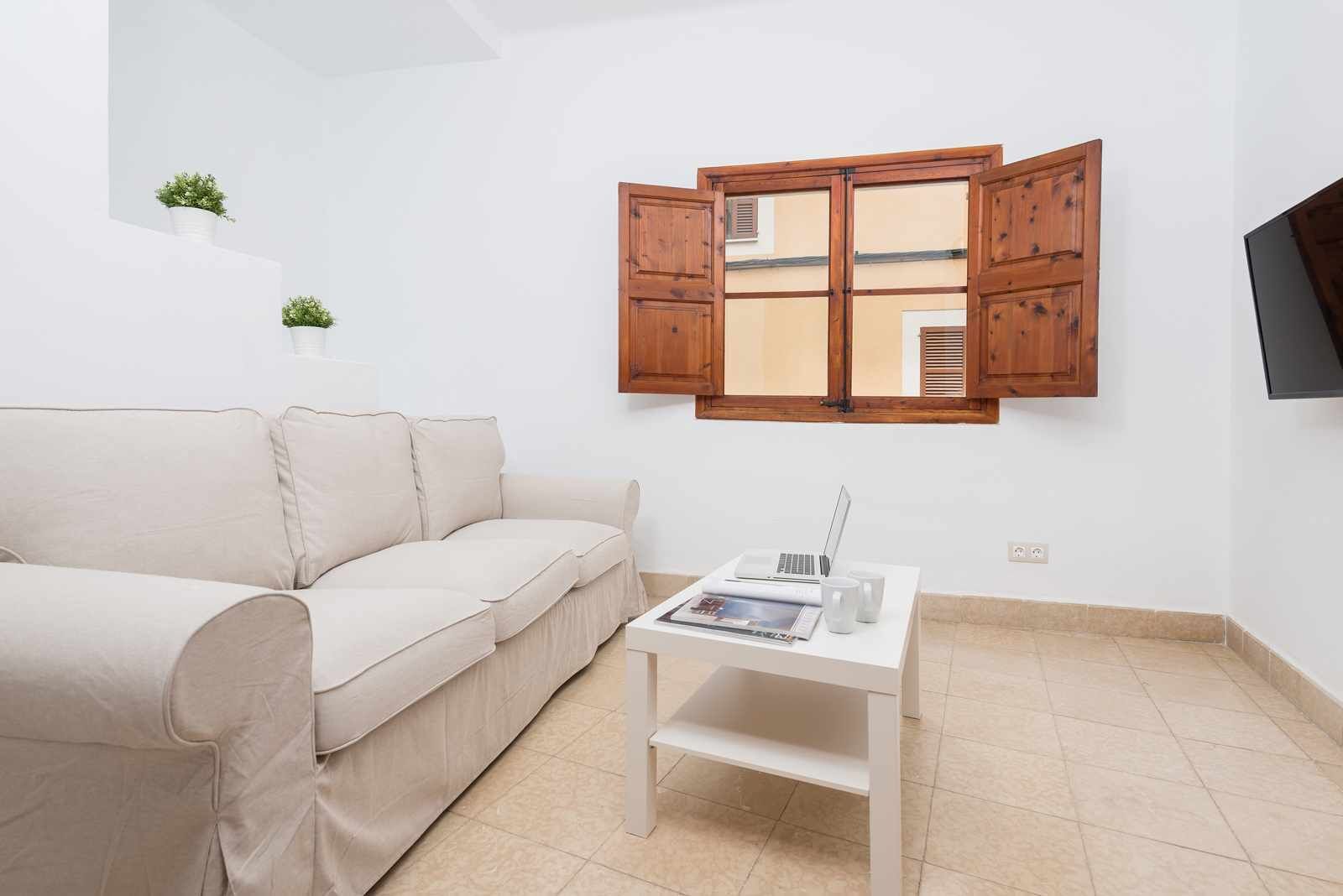 Een minimalistische woonkamer met een beige bank, een klein wit tafeltje met een laptop en een tijdschrift, en een houten kast met een raam erboven.
