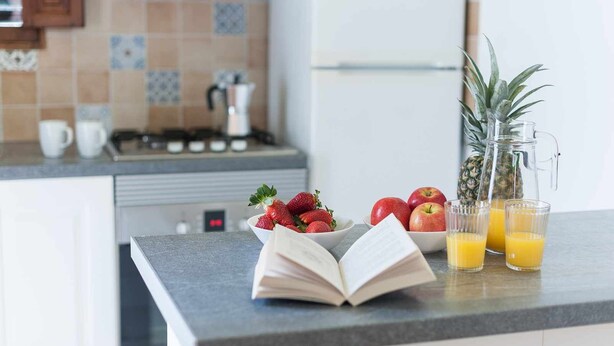 Una encimera de cocina con un libro, un cuenco de fresas, un cuenco de manzanas, una piña y dos vasos de zumo de naranja.