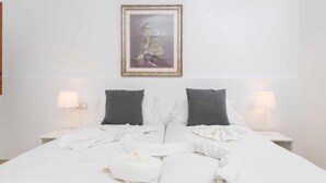 Una cama bien hecha con sábanas blancas y dos almohadas oscuras, un retrato enmarcado en la pared y dos lámparas de noche a cada lado.