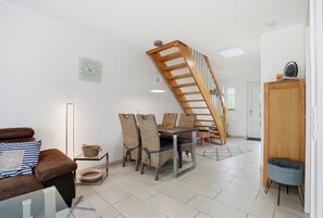 A modern living room with a wooden staircase, a dining area with wicker chairs, a brown leather sofa, and a small round table with a basket.