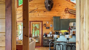 A rustic cabin interior with a wooden ceiling, a mounted animal head, a kitchen area with bar stools, and a cozy seating area. - Experience North GA Mtn Retreat: Views w/ Hot Tub (Morganton)