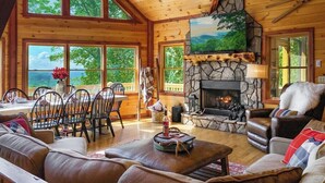 A log cabin interior with a stone fireplace, wooden dining area, and large windows offering a view of the outdoors. - Experience North GA Mtn Retreat: Views w/ Hot Tub (Morganton)
