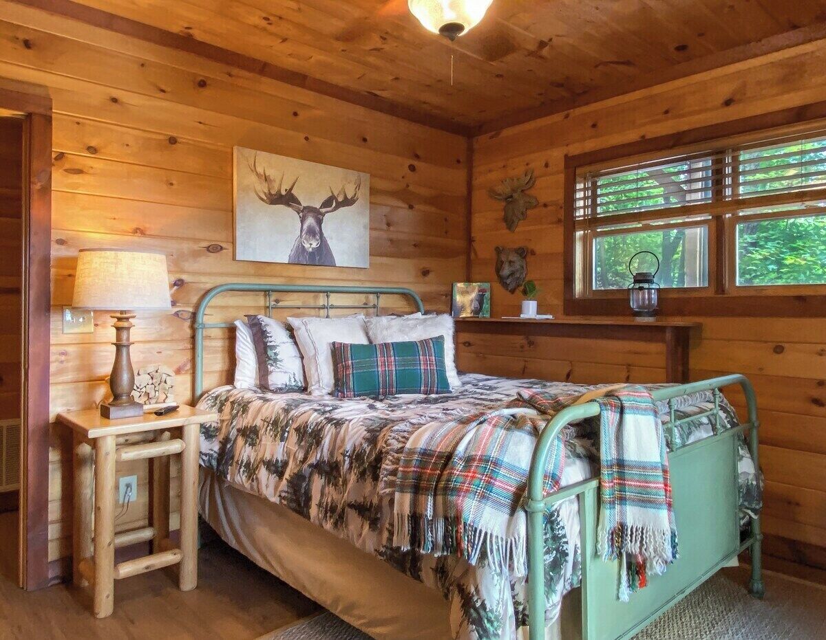 A wooden cabin bedroom with a metal bed, a wooden nightstand, a lamp, a framed moose head, and a window with a view of trees.