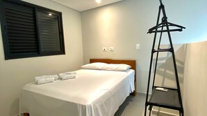 A bedroom with a bed, a window with blinds, a bedside table, and a black shelf unit. - Duplex apartment in Villa Licuri (são sebastião)
