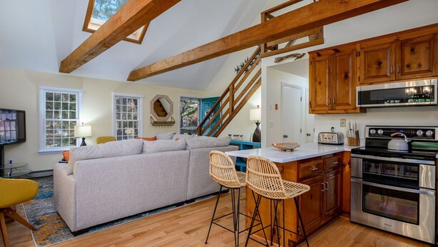 Eine moderne Küche mit Holzausstattung, einem Einbauofen und einer Mikrowelle. Es gibt eine Essecke mit Barhocker und einer Couch.