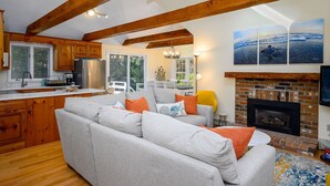 Ein gemütliches Wohnzimmer mit grauem Sofa, Holzbalken, Kamin und einer Küchenzeile mit Holzausbau und einem Fenster.