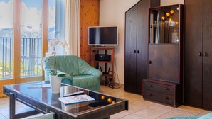 Una habitación con una mesa de vidrio, una silla verde, un televisor sobre un mueble bajo, un armario y vistas a montañas a través de amplios ventanales.