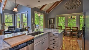 A modern kitchen with a built-in dishwasher, white cabinetry, and a large island with a sink. There are large windows providing natural light and views of greenery outside.