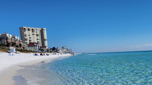 A beach with clear turquoise water, a sandy shore, and buildings in the background.