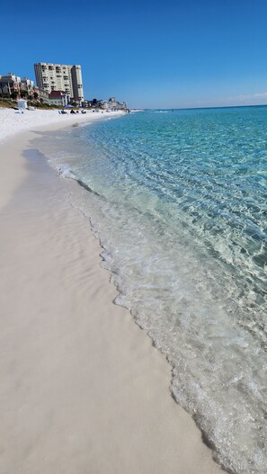 A beach with clear turquoise water, a sandy shore, and buildings in the background.