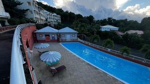 Ein Swimmingpool mit Liegestühlen und Sonnenschirmen, umgeben von einem Zaun, mit Gebäuden im Hintergrund.