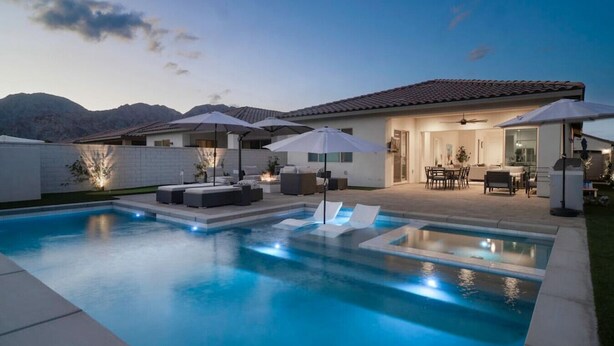 A modern outdoor pool area with lounge chairs and umbrellas, a patio with a dining set, and a view of mountains in the background. - Private pool & hot tub w/ mountain views (La Quinta)