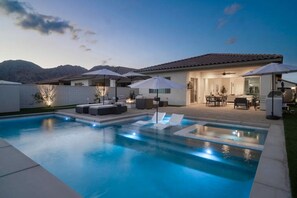 A modern outdoor pool area with lounge chairs and umbrellas, a patio with a dining set, and a view of mountains in the background. - Private pool & hot tub w/ mountain views (La Quinta)