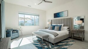 A modern bedroom with a large bed, a ceiling fan, a window with a view, and a nightstand with a lamp. - Private pool & hot tub w/ mountain views (La Quinta)