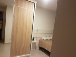 A bedroom with a wooden wardrobe, a bedside table, and a bed with a beige cover. - Apartment across from Praia Grande beach with an excellent view/unparalleled sunset! (Arraial do Cabo)