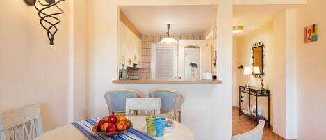 A dining area with a round table, chairs, and a bowl of fruit. There is a kitchen visible in the background with a white door and a mirror above the counter.
