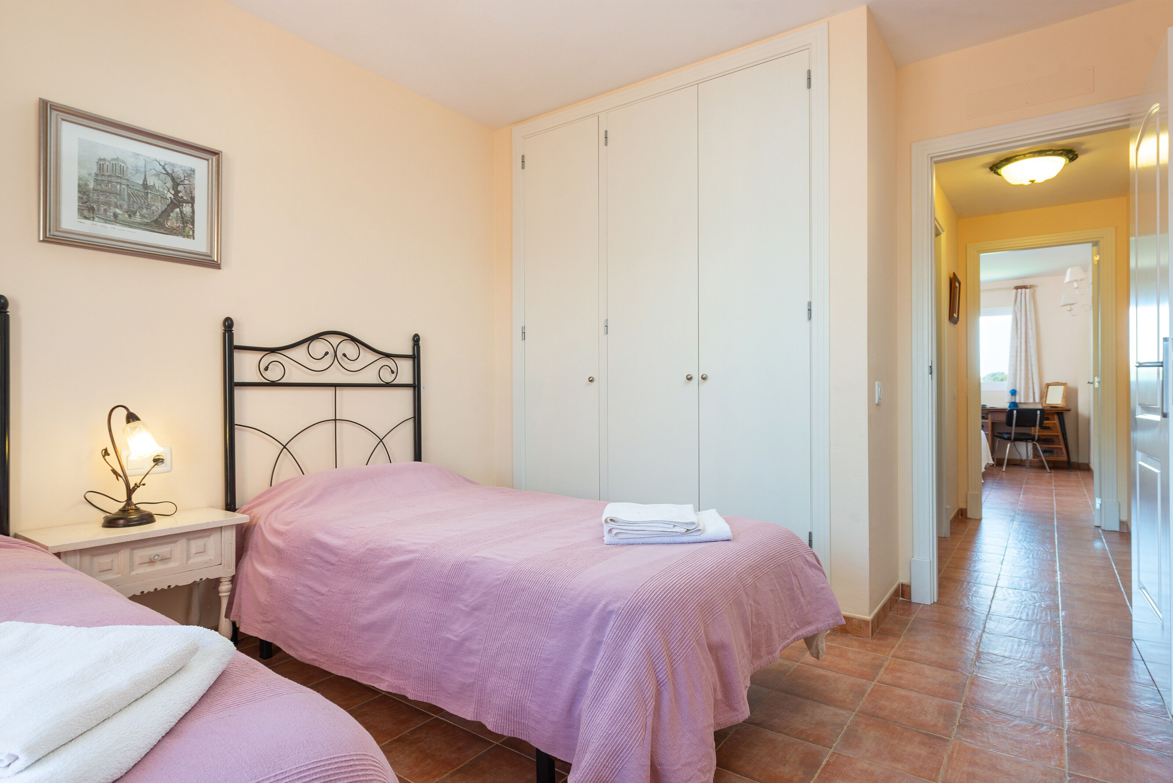 A bedroom with two beds, a nightstand with a lamp, a framed picture on the wall, and a white wardrobe.