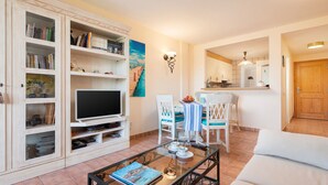 Una sala de estar con televisor, estanterías, un comedor con sillas y mesa, y una cocina al fondo.