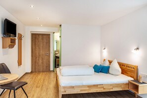 A bedroom with a wooden bed, a TV mounted on the wall, a desk with a chair, and a door leading to another room.