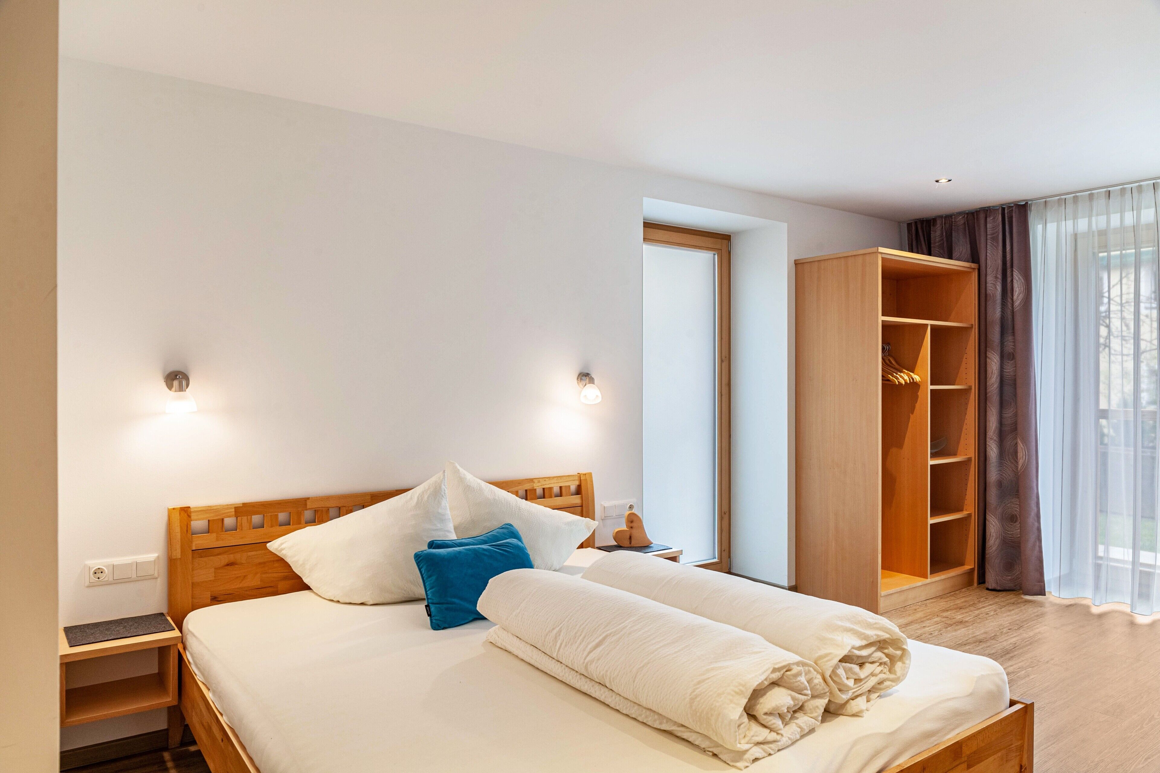 Una cama bien hecha con almohadas, una mesita de noche de madera, un armario y una ventana con cortinas.