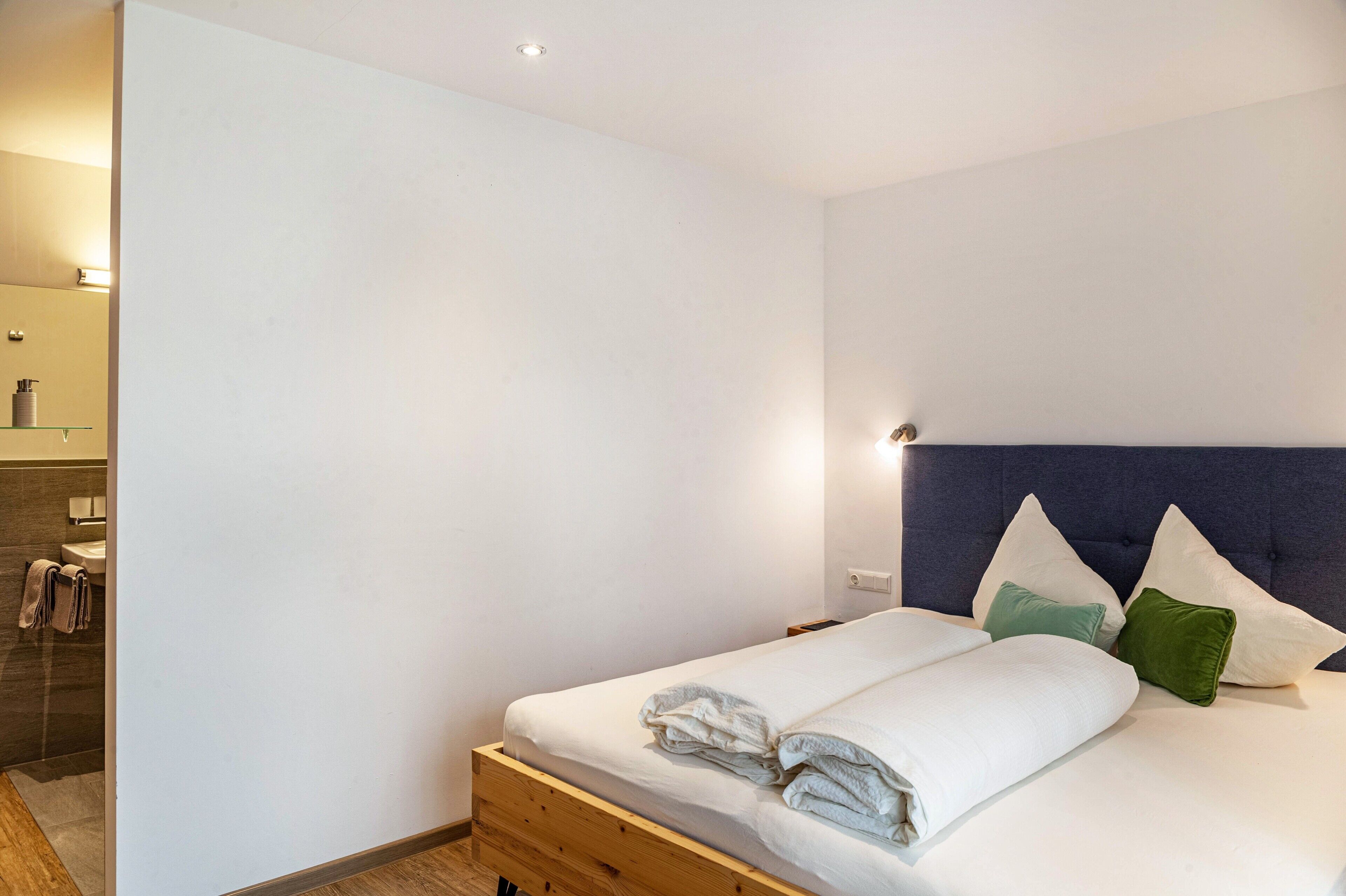 A bedroom with a bed, a nightstand, a wall-mounted light, and a bathroom with a mirror and towel rack.