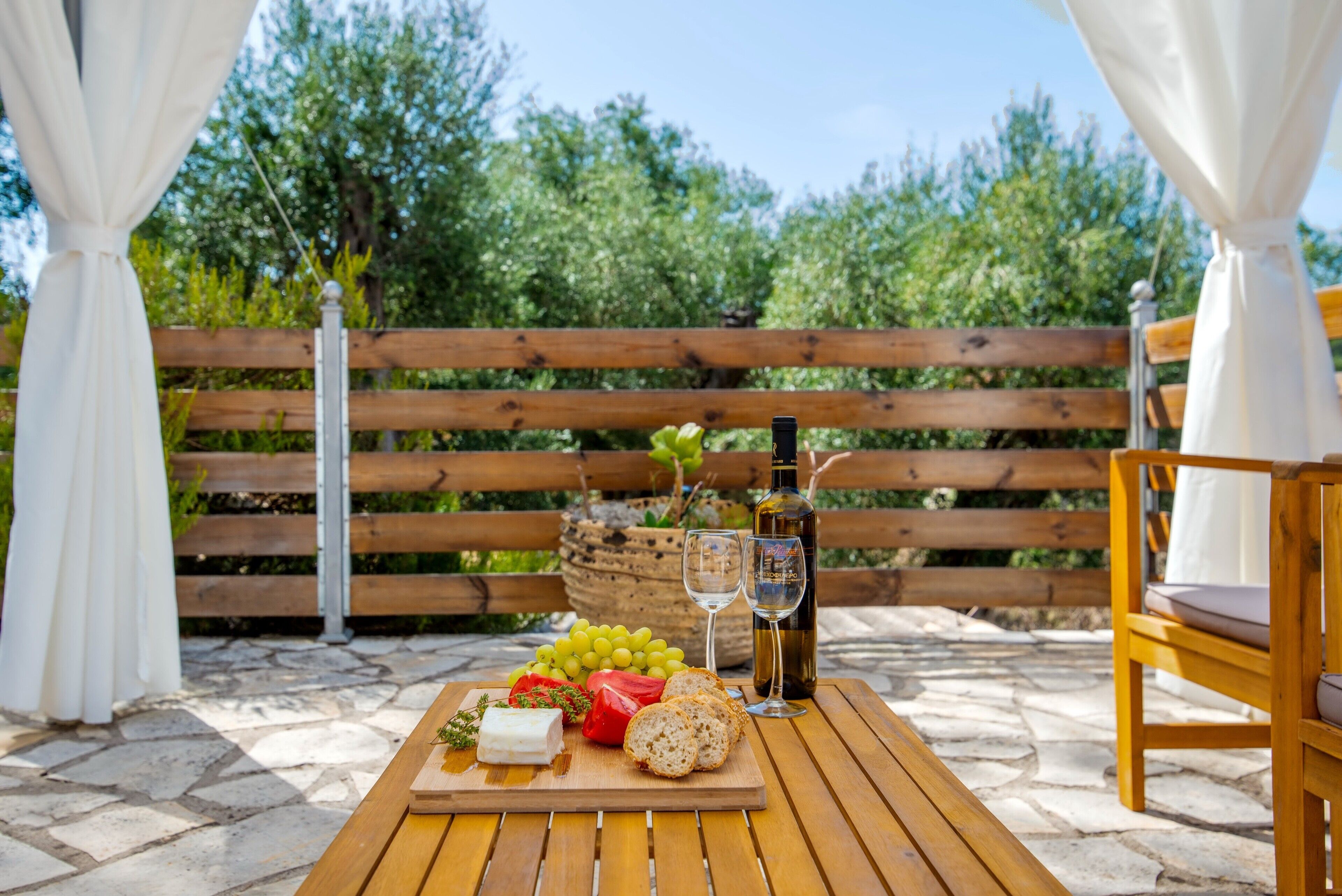 A wooden table with a cheese board, grapes, a bottle of wine, and a glass on a patio with a wooden bench and white curtains.