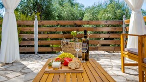 A wooden table with a cheese board, grapes, a bottle of wine, and a glass on a patio with a wooden bench and white curtains. - Holiday Home 'Ria Sweet Home Paxos' with Private Terrace, Private Garden and Wi-Fi (Corfu)