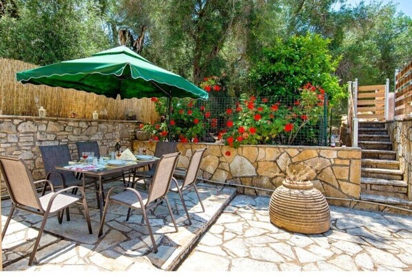 An outdoor patio with stone flooring, a table with chairs, a green umbrella, and a stone wall with red flowers.