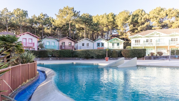 A swimming pool with a curved edge, surrounded by colorful houses with balconies, and a forested background. - House with View, Shared Pool and Garden (Lacanau-Ocean)