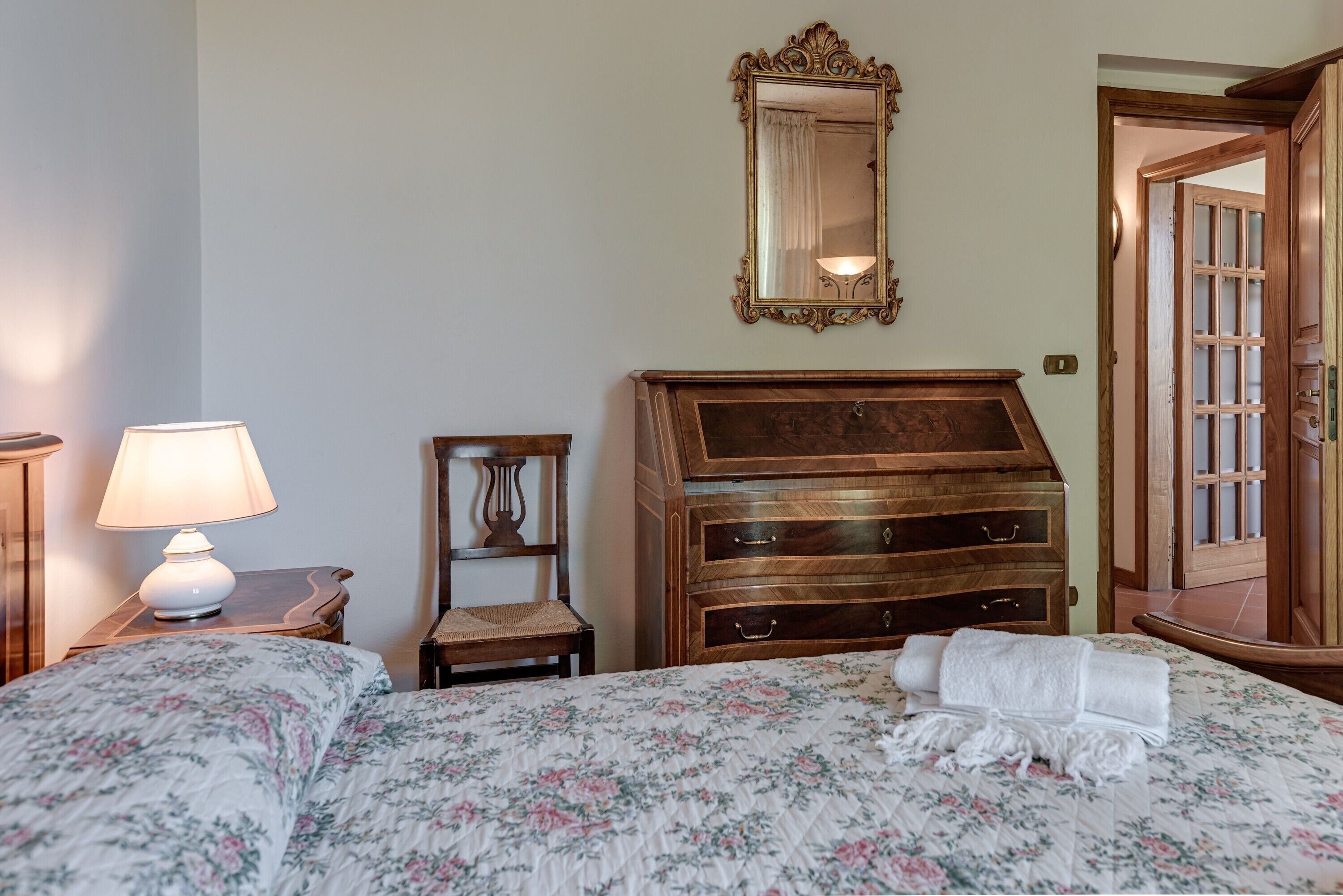 A bedroom with a bed, a dresser, a chair, a lamp, a mirror, and a door.
