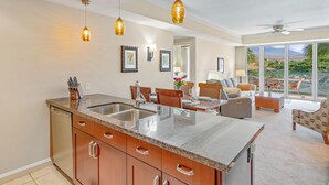 A modern kitchen with granite countertops, a built-in sink, and wooden cabinets. There is a dining area with a table and chairs, and a living room with a sofa and a coffee table. The room has large windows offering a view of the outdoors. - Maui Resort Rentals: Honua Kai Konea 308 - Spacious  2BR/1BA, Gorgeous Partial Ocean and Lush West Maui Mountain Views! (Lahaina)