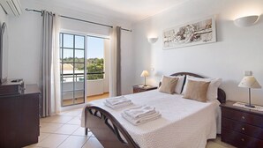 A bedroom with a bed, bedside tables, a nightstand, a window with curtains, a wall-mounted artwork, and a view of greenery outside. - "Villa Catarina" Vacation Home with Pool & Wi-Fi (Albufeira)