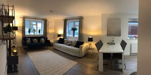 En moderne stue med sofa, spisebord og sofabord. To vinduer med gardiner, en plante og en lampe.