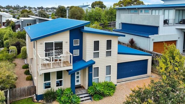 Une maison moderne à deux étages, avec un toit bleu et des murs de couleur beige, dotée d’un balcon et d’un garage.