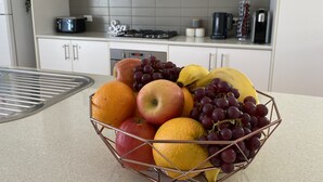 Une cuisine avec des armoires blanches, un évier en acier inoxydable et un panier de fruits contenant des pommes, des oranges, des bananes et des raisins, posé sur le comptoir.
