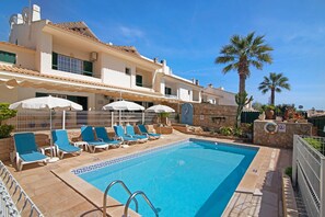 Una piscina con sillones de descanso y sombrillas, un muro de piedra y palmeras.