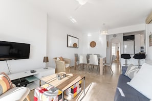 Un salon moderne avec un téléviseur à écran plat, une table basse en bois, un canapé, un coin repas avec des chaises et une cuisine en arrière-plan.