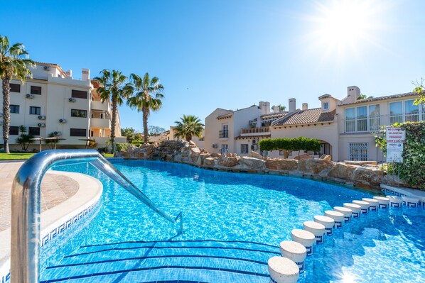 A swimming pool with a stone border, palm trees, and buildings in the background.
