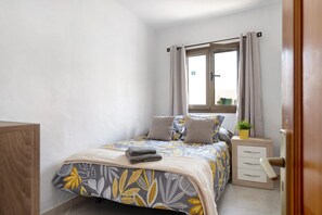 A bedroom with a bed, a bedside table with a plant, a window with curtains, and a door.
