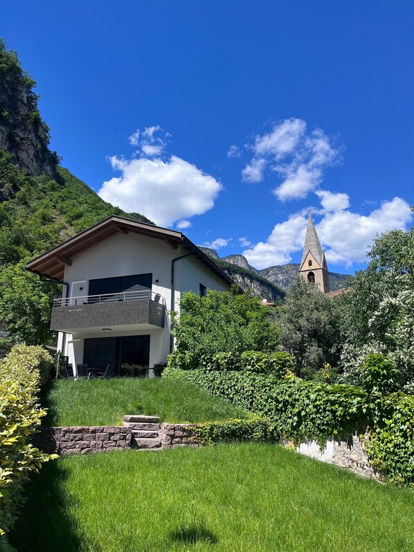 Una casa moderna de dos pisos con un balcón, rodeada de exuberante vegetación y con montañas al fondo.
