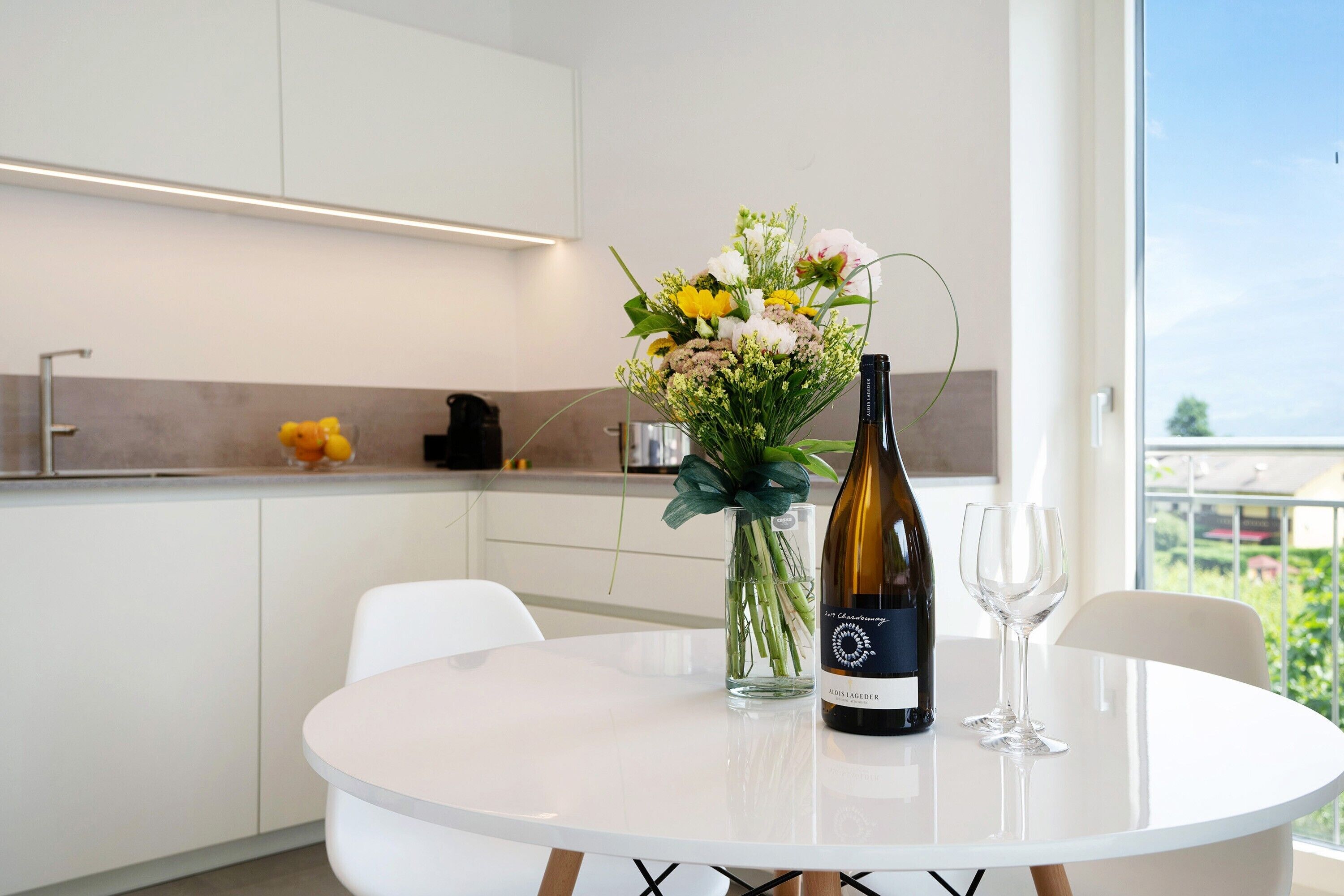 Une cuisine moderne avec des meubles blancs, une table ronde blanche, une bouteille de vin et un vase de fleurs.