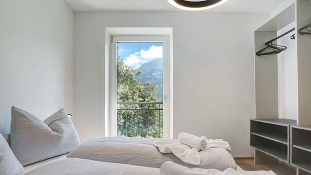 Een slaapkamer met een bed, een raam met uitzicht op groen en bergen, een kledingkast en een hangrek.