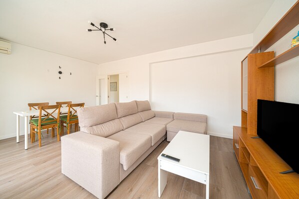 Ein modernes Wohnzimmer mit einer Eckcouch, einem Esstisch mit Stühlen, einer TV-Konsole und einer Deckenleuchte.