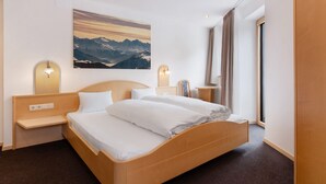Une chambre d’hôtel avec un lit en bois, une table de chevet, une lampe fixée au mur, un tableau représentant des montagnes et une fenêtre avec des rideaux.
